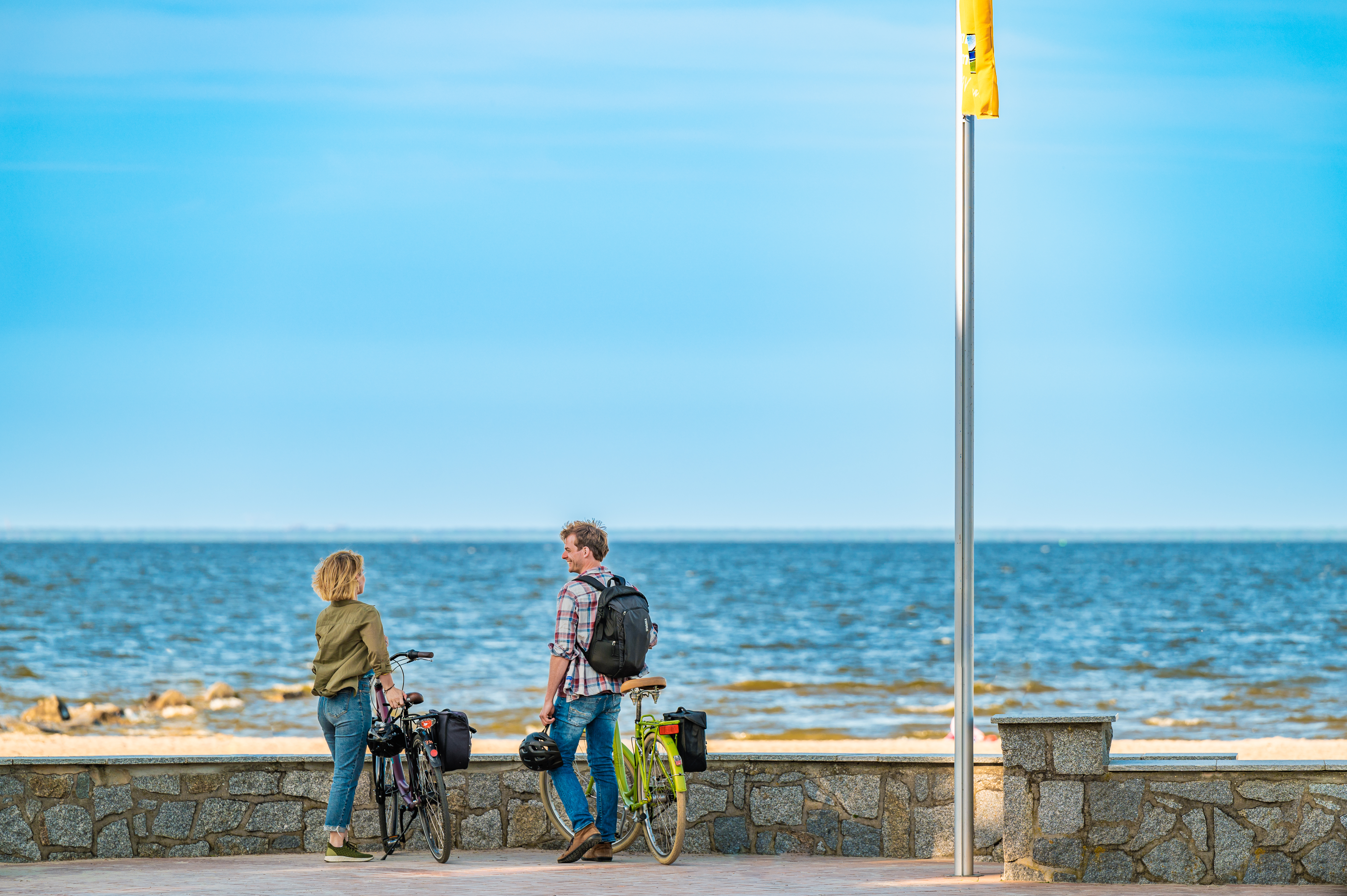 Ein Paar steht mit seinen Fahrrädern vor einer kleinen Steinmauer und blickt aufs Meer. Neben ihnen ist ein Fahnenmast und daran hängt eine gelbe Fahne